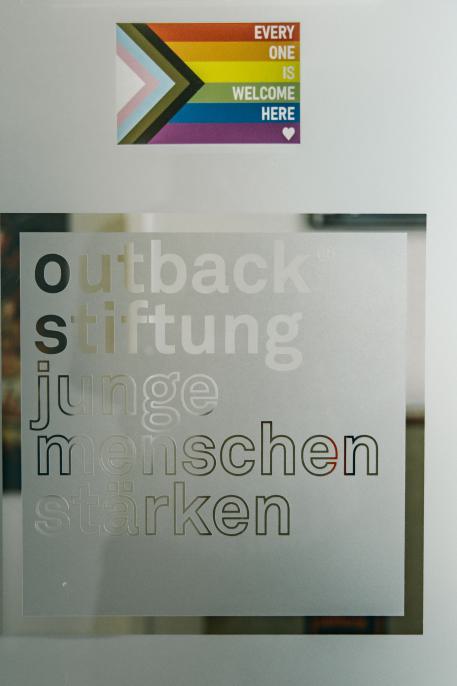 Selbstständigkeit in der Jugendhilfe, wozu? Durchsichtige Tür it dem outback Logo drauf, darüber ein Sticker von der progressiven Pride Flagge
